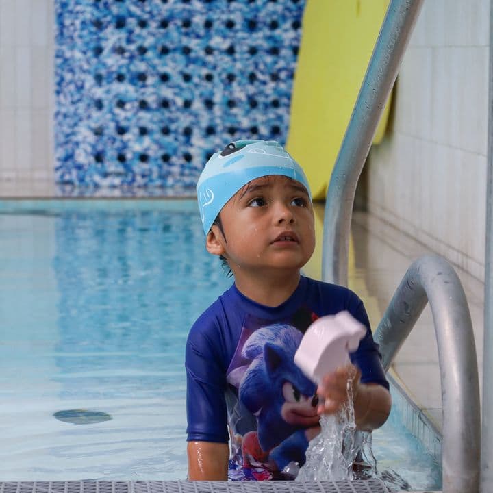 Un niño con su gorro azul se apoya en la escalera de la alberca