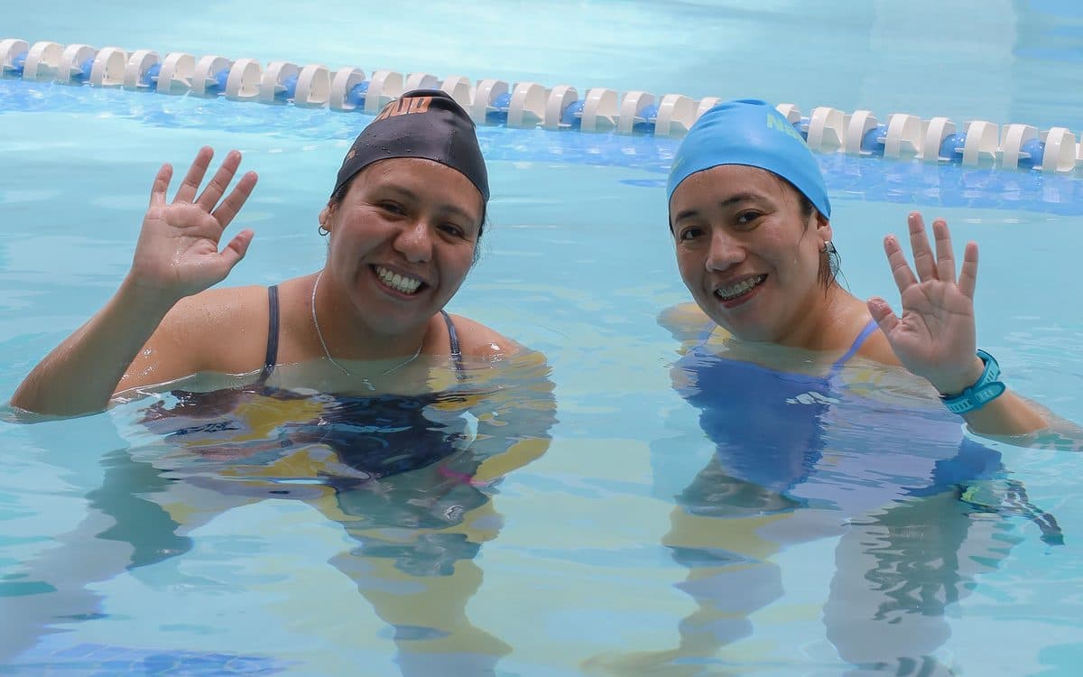 Dos alumnas adultas sonríen y saludan desde la alberca